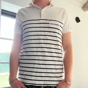 Hilfiger piké  - Säljer en vit piké från Hilfiger med svarta horisontella ränder och en liten logga på bröstet. T-shirten har korta ärmar och klassisk krage. Perfekt för en stilren och avslappnad look.