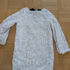 Vit paljettklänning med svart rosett stl xs Ny - Säljer en vit glittrig klänning täckt av paljetter med trekvartsärm och en stor svart rosett i nacken. Klänningen har en rak passform och är perfekt för dig som vill sticka ut. Endast provad ny