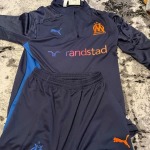 Marinblå långärmad fotbollströja från Puma, Olympique de Marseille - Säljer en marinblå långärmad fotbollströja från Puma med Olympique de Marseille-logga och orange detaljer. Tröjan har halv dragkedja och tryckta loggor på bröstet. Matchande byxor medföljer, båda med blå och orange detaljer.