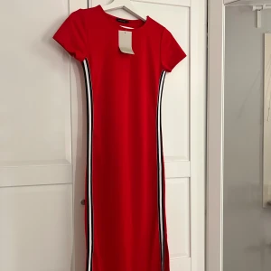 Röd klänning med ränder från DeFacto - Säljer en röd kortärmad klänning från DeFacto med svarta och vita ränder längs sidorna. Klänningen har rund halsringning och en figurnära passform. Perfekt för dig som gillar en enkel men snygg stil.