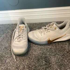 Vita Nike Kobe sneakers med gulddetaljer - Säljer ett par vita Nike Kobe sneakers med guldfärgade detaljer och logga. Skorna har ett mönstrat mesh-överdrag och klassisk snörning. Perfekta för dig som gillar basket eller vill ha en snygg streetstil.TVÄTTAR sulan före lämning⚠️