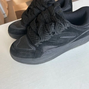Svarta sneakers med grova snören - Säljer ett par helsvarta sneakers med riktigt breda och grova snören. Skorna har en sportig look med rund tå och platt sula. Passar perfekt till dig som vill ha en unik detalj på fötterna. Materialet är syntet och textil.