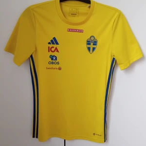Svenska landslagets fotbollströja från Adidas - Gul kortärmad fotbollströja från Adidas med blå detaljer och svenska landslagets emblem på bröstet. Tröjan har tryckta sponsorer på framsidan och blå ränder längs sidorna. Tillverkad i lätt och ventilerande material.