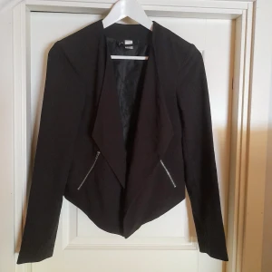 Svart kavaj från H&M Divided  - Snygg svart kavaj från Divided i storlek 36. Jackan har lång ärm, öppen front och två sneda dragkedjor framtill. Materialet är en mix av polyester, viskos och elastan vilket ger en skön passform. Perfekt för en stilren look. Använd en gång 