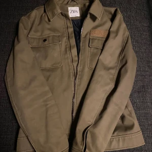 Olivgrön jacka från Zara - Säljer en olivgrön jacka från Zara med klassisk krage och två bröstfickor med lock. Jackan har synliga sömmar och stängs med dragkedja. Perfekt för lager-på-lager och enkel att matcha med olika outfits.