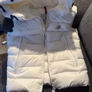 Vit väst Moncler - Säljer en snygg vit dunväst från Moncler med huva och dragkedja framtill. Västen har två fickor med dragkedja. Den är I mycket fint skick 
