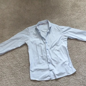 Ljusblå skjorta från Eton - Säljer en stilren ljusblå skjorta från Eton i slim fit-modell. Skjortan har klassisk krage, långa ärmar och knäppning framtill. Perfekt för dig som gillar en enkel och snygg look. Materialet är tunt, mjukt och svalt.