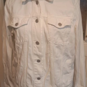 Vit jeansjacka från GAP - Säljer en klassisk vit jeansjacka från GAP i storlek L. Jackan har långa ärmar, knappar framtill och två bröstfickor med lock och knapp. Perfekt att slänga över en t-shirt för en stilren look.