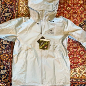 Arc'teryx - Ljusgrå - Säljer en stilren ljusgrå skaljacka från Arc'teryx i GORE-TEX-material. Jackan har en justerbar huva och flera praktiska dragkedjefickor. Perfekt för regniga dagar och utomhusaktiviteter.