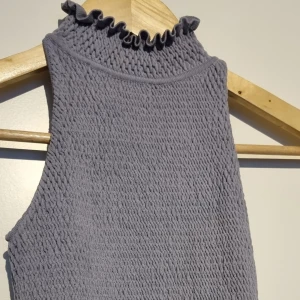 Grå smockad ärmlös topp från Zara - Supersöt ljusgrå ärmlös topp från Zara med smockat tyg och volangdetaljer vid hals och nederkant. Perfekt att styla till jeans eller kjol för en trendig look!