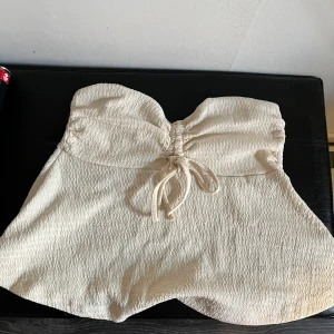 Gul axelbandslös tröja med knyt - Säljer en söt axelbandslös tröja i gul med struktur. Tröjan har en snygg knytning framtill. Perfekt för varma dagar eller sommarens tillfällen.