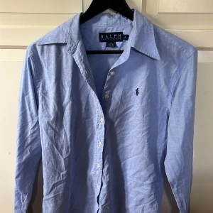 Ljusblå skjorta från Ralph Lauren - Klassisk ljusblå skjorta från Ralph Lauren med broderad logga på bröstet. Skjortan har lång ärm, knappar framtill och är tillverkad i 100% bomull. Perfekt för en stilren och tidlös look.