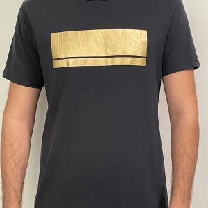 Marinblå t-shirt med guldtryck från Hugo Boss - Snygg marinblå t-shirt från Hugo Boss med stort guldigt BOSS-tryck på bröstet. Klassisk passform och rund halsringning. Tröjan är använd ett par gånger bara men är till salu då den inte kommer till användning. T shirtens storlek är i L men har passform för M! 