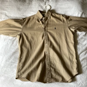 Beige skjorta från Ralph Lauren - Klassisk beige skjorta från Ralph Lauren med blå broderad logga på bröstet. Skjortan har lång ärm, knappar framtill och är tillverkad i 100% bomull. Perfekt för en stilren och tidlös look. Den är väldigt stor i storleken även fast det står small. Men hör av dig så fixar jag mått 👍 