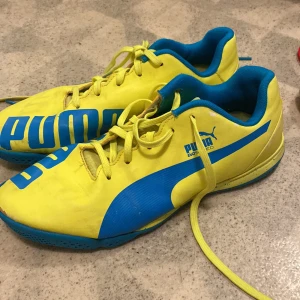 Gula och blå fotbollsskor från Puma - Säljer ett par gula och blå fotbollsskor från Puma med snörning och stor logga på sidan och tån. Skorna har platt sula och är perfekta för inomhusspel. Snygg sportig design och bekväm passform.
