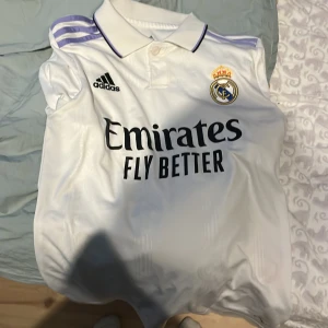 Real Madrid hemmatröja Adidas - Vit Real Madrid fotbollströja från Adidas med klubbmärke och sponsortryck på bröstet. Tröjan har lila detaljer på axlarna och klassisk krage med knappar. Perfekt för dig som vill visa ditt stöd för laget.