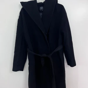 Svart kappa med bälte från Dixi Coat - Säljer en svart lång kappa från Dixi Coat med bälte i midjan och stor krage. Kappan har lång ärm och är fodrad på insidan. Perfekt för kyligare dagar och ger en stilren look.