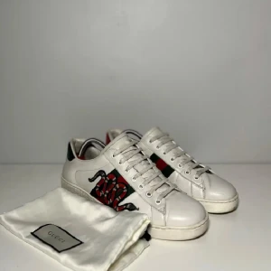 Gucci vita sneakers med orm brodyr - Säljer ett par vita sneakers från Gucci med ikonisk röd och grön rand samt broderad orm på sidan. Skorna har rund tå, platt sula och detaljer i rött och grönt på hälen. Klassisk och stilren design med snörning framtill.