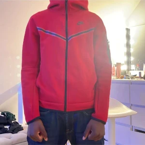 Röd Nike hoodiejacka med dragkedja - Säljer en röd hoodiejacka från Nike med hel dragkedja framtill och svarta detaljer. Jackan har huva, lång ärm och en sportig design med svarta linjer över bröstet. Perfekt för dig som gillar streetwear och sportig stil.