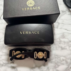 Svarta solglasögon från Versace - Snygga svarta solglasögon från Versace med rektangulära bågar och guldiga detaljer på sidorna. Klassisk design med mörka glas och exklusiv känsla. Kommer med originaletui och ask.