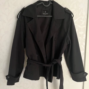 Svart trenchcoat från Stradivarius - Snygg svart trenchcoat från Stradivarius med bälte i midjan och knappar framtill. 