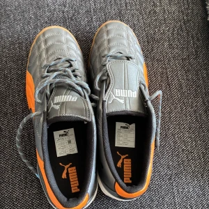 Gråa och orangea fotbollsskor från Puma - Säljer ett par gråa fotbollsskor från Puma med orangea detaljer och vit sula. Skorna har snörning och är designade med rund tå och platt sula. Perfekta för dig som vill sticka ut på planen!