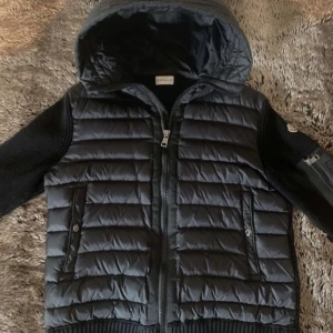 Svart pufferjacka med stickade ärmar från Moncler - Säljer en svart pufferjacka från Moncler med stickade ärmar och huva. Jackan har dragkedja framtill, två fickor med knappar och en extra ficka på ärmen. Perfekt för kyliga dagar och riktigt snygg streetstil. Storlek L men kan passa som M, vid snabb affär kan jag gå ner i pris