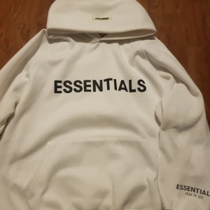 Vit hoodie från Essentials Fear of God - Säljer en vit hoodie från Essentials Fear of God med svart tryck på bröstet och ärmen. Klassisk känguruficka framtill och huva. Perfekt för en clean och stilren look.