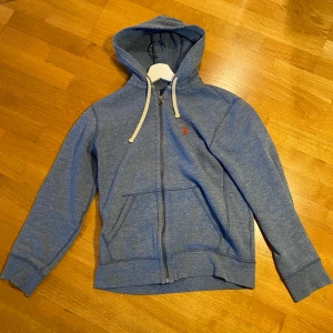 Blå hoodie från Polo Ralph Lauren - Säljer en blå hoodie från Polo Ralph Lauren med dragkedja och vit dragsko. Tröjan har fickor framtill och en liten röd logga broderad på bröstet. Perfekt för en avslappnad stil.