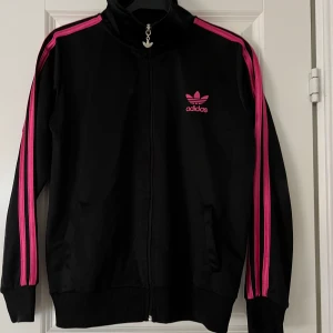 Svart träningsjacka Adidas - Snygg svart träningsjacka från Adidas med rosa detaljer och logga. Strl. 170-176, i mycket bra skick. Jackan har dragkedja framtill och långa ärmar med rosa ränder längs sidorna. 