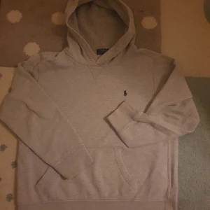 Grå hoodie från Polo Ralph Lauren - Säljer en stilren grå hoodie från Polo Ralph Lauren med klassisk broderad logga på bröstet. Tröjan har huva, magficka och långa ärmar. Perfekt för en avslappnad och snygg look.