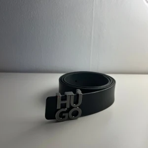 Svart bälte från Hugo med metallspänne - Snyggt svart bälte från Hugo med ett stilrent metallspänne där det står 'HUGO' i stora bokstäver. Bältet är gjort i slätt skinn och passar perfekt till jeans eller kostymbyxor. Enkel och modern design som lyfter din outfit.