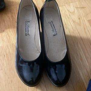 Svarta lackade wedges från E. Shamel - Säljer ett par svarta glansiga wedges med rund tå från E. Shamel. Skorna har hög kilklack och platt sula undertill. Perfekta för dig som vill ha lite extra höjd men ändå bekvämt.