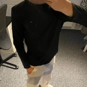 Svart långärmad tröja Tommy Hilfiger - Svart långärmad tröja från Tommy Hilfiger med klassisk rund hals och liten broderad logga på bröstet. Tillverkad i mjuk bomull som känns skön mot huden. Perfekt för en clean och stilren look.