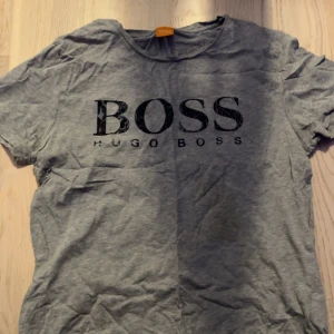 Grå t-shirt från Hugo Boss - Snygg grå t-shirt från Hugo Boss med stor svart logga på bröstet. Klassisk rund halsringning och korta ärmar. Perfekt basic-plagg med stilren design och skön passform. Materialet känns mjukt och passar till många olika outfits.