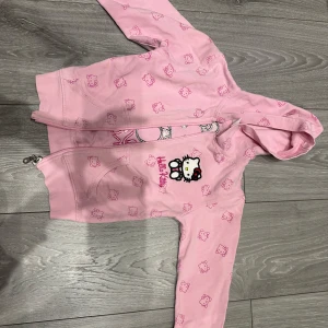 Hello Kitty rosa hoodie med dragkedja för barn  - Supersöt rosa hoodie med dragkedja och huva från Hello Kitty. Jackan har ett all-over Hello Kitty-mönster och en stor broderad Hello Kitty-figur framtill tillsammans med texten 'Hello Kitty' i rosa. Perfekt för dig som älskar gulliga detaljer och vill sticka ut. Men dragkedjan fungerar inte riktigt. 