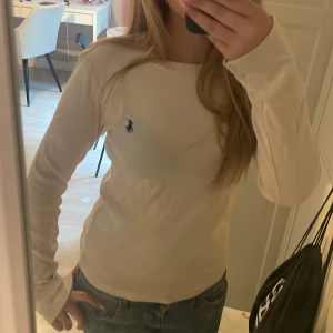 kräm vit långärmad tröja från Ralph Lauren - Säljer en stilren kräm vit långärmad tröja från Ralph Lauren med den klassiska blå loggan broderad på bröstet. Tröjan är i mjuk bomull och har en slim passform som sitter snyggt. Perfekt till jeans för en clean och enkel look.