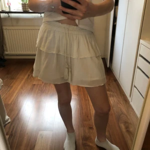 Vit volangkjol från Gina Tricot - Superfin vit kortkjol från Gina Tricot med volanger och knytband med pärldetaljer och tofsar i midjan. Kjolen är i ett lätt och luftigt material, perfekt för varma dagar. Sitter skönt med resår i midjan