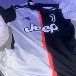 Juventus Adidas fotbollströja S - Juventus matchtröja från Adidas i storlek S. Tröjan har svart och vit färgblock med en röd linje i mitten, Juventus-logga med tre stjärnor, Adidas-märke och Jeep som sponsor. Tillverkad i lätt och ventilerande Climalite-material, perfekt för fotbollsträning eller match.