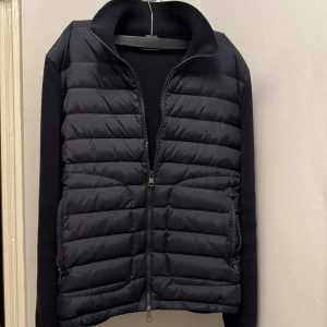 Moncler Cardigan - Snygg marinblå cardigan från Moncler med quiltad framsida och stickade ärmar. Västen har hög krage, dragkedja framtill och två dragkedjefickor. Klassisk Moncler-logga på ärmen. Perfekt för lager-på-lager och stilren look. Bara höra av sig om minsta lilla😊