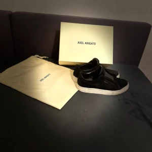 Svarta sneakers från Axel Arigato - Stilrena svarta sneakers från Axel Arigato i mocka med vit platt sula. Klassisk låg modell med diskret logga på sidan och elastisk detalj över vristen. Perfekta för dig som gillar minimalistisk och modern design. Skorna är i helt okej skick använda i ett ett tag säljer de pga av att har blivit för små 