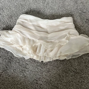 Vit volangkjol i sidenlook - Supersöt kort vit kjol med flera lager volanger och glansig sidenkänsla. Kjolen har en flowy och luftig stil som ger en riktigt romantisk vibe. Perfekt för dig som vill sticka ut med en feminin och trendig look. Inbyggda shorts i 