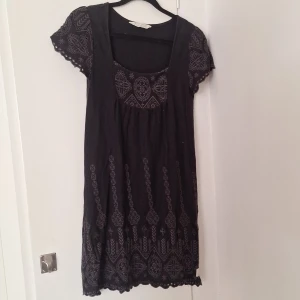 Svart kortklänning Odd Molly med broderi - Supersöt svart kortklänning från Odd Molly med grått broderat mönster och korta ärmar. Klänningen har rundad halsringning och fina detaljer längs fållen och ärmsluten. Perfekt för dig som gillar boho-vibe och snygga broderier.