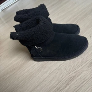 Svarta boots med fuskpäls och spänne - Mysiga svarta boots med mjukt foder i fuskpäls och snygga spänndetaljer på sidan. Skorna har rund tå, platt sula och är tillverkade i mocka-liknande material. Perfekta för kalla dagar när du vill hålla stilen och värmen.