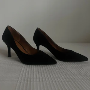 Svarta pumps  - Klassiska svarta pumps i mocka med spetsig tå och smal klack. Höjden på klacken är cirka 6-7 cm. Som bilden visar finns det lite slitningar på klacken annars är dom felfria.