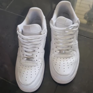 Nike Air Force 1 helvita sneakers - Klassiska Nike Air Force 1 sneakers i helvitt skinn med perforerad tå, rund tå och platt sula. Swoosh-logga på sidan och snörning framtill. Perfekta för dig som gillar en clean och tidlös look.