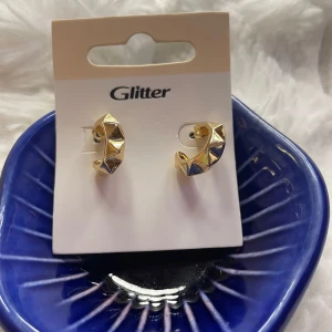 Guldiga örhängen från Glitter - Snygga Stockholms stilla örhängen i guld. Coola guldiga örhängen från Glitter med chunky halvcirkel-form och pyramidformade detaljer. De har en modern och edgy vibe som lyfter vilken outfit som helst. Perfekta för dig som gillar statement-smycken och vill sticka ut lite extra. Helt nya med prislapp 