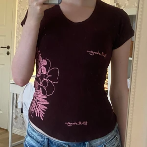 Brun t-shirt med blommor från Billabong - Snygg brun t-shirt från Billabong med rosa blommigt tryck på sidan och logga framtill. Modellen har v-ringning och korta ärmar, sitter figurnära och är gjord i mjuk bomull. Perfekt för dig som gillar surf- och streetstil.