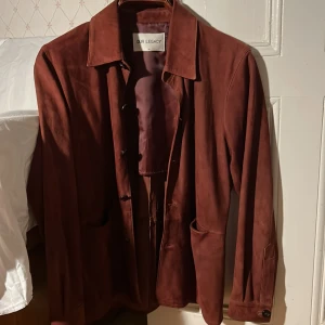 Brun mocka overshirt från Our Legacy - Säljer en brun overshirt i mjuk mocka från Our Legacy. Jackan har klassisk krage, knäppning framtill och två stora fickor. Perfekt för lager-på-lager och ger en clean och stilren vibe. Passar dig som gillar minimalistisk och tidlös design.