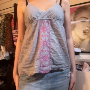 Grå linnetopp med broderi - Supersöt grå linnetopp med tunna axelband och v-ringning. Framtill finns ett rosa broderat mönster som ger toppen en unik touch. Luftig och perfekt för varma dagar, lätt att matcha med jeans eller kjol.